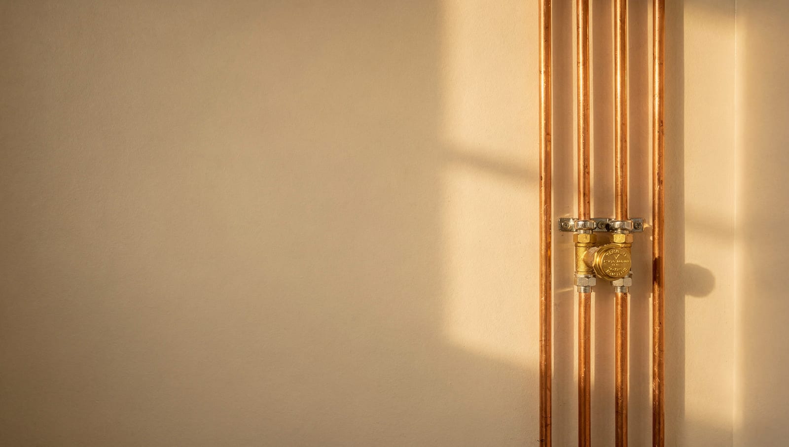 Neatly run copper supply lines and a brass shutoff valve on a warm cream wall in golden light