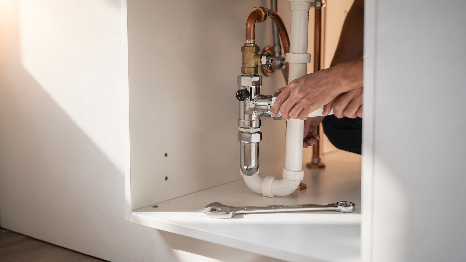 A plumber's hand on a polished shutoff valve beside clean copper supply lines under a sink